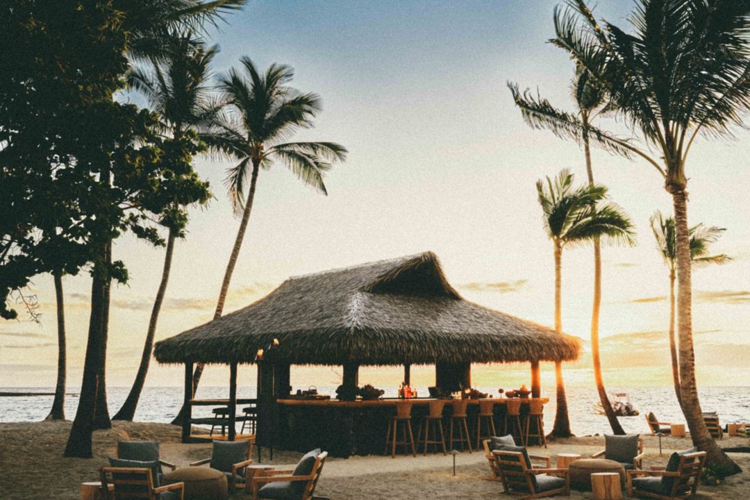 Beach bar with a thatched roof, lounge seating, and a sunset ocean backdrop at a high-end Kona resort.